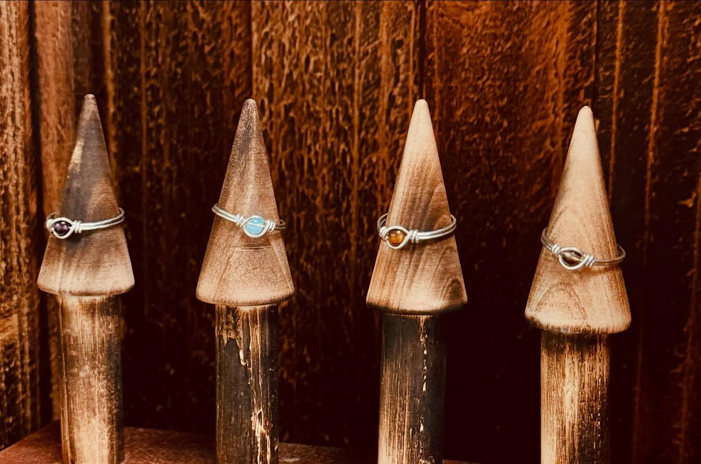 Four silver rings with different colored gemstones displayed on pointed wooden stands against a dark wood background.